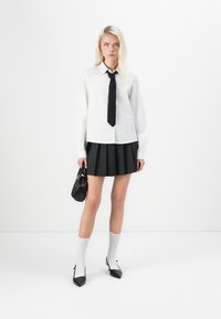 Weißes Langarmhemd mit einer schwarzen Krawatte, kombiniert mit einem schwarzen, plissierten Minirock. Schwarze High-Heels und knielange weiße Socken. Trägt eine kleine schwarze Tasche.