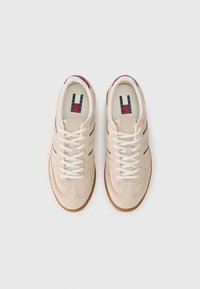 Μπεζ σουέντ αθλητικά παπούτσια με λευκά κορδόνια, ρίγες μπλε λεπτομέρειες και λάστιχο από καουτσούκ. Η ετικέτα με το λογότυπο στην γλώσσα διαθέτει κόκκινα, λευκά και μπλε χρώματα.