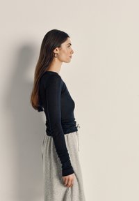 Mlada žena s dugom tamnom kosom, odjevena u crnu majicu dugih rukava i sivu suknju, stoji u profilu ispred jednobojnog svijetlog zida.