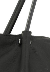 Dettaglio di una borsa in pelle nera che mostra la superficie testurizzata, i bordi cuciti e gli accenti in metallo argentato sulla tracolla e sul corpo della borsa.