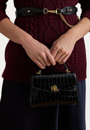 Melnas krokodila rakstā iegremdētas rokas somas ar zelta logo detaļu, ar strukturētu formu un augšējo rokturi, turēta pretī bordo adītai džemperim.