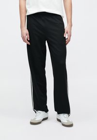 Μαύρο αθλητικό παντελόνι με υφή υφάσματος, με λευκές παράλληλες ρίγες στο πλάι και άνετη εφαρμογή, συνδυασμένο με λευκά αθλητικά παπούτσια.