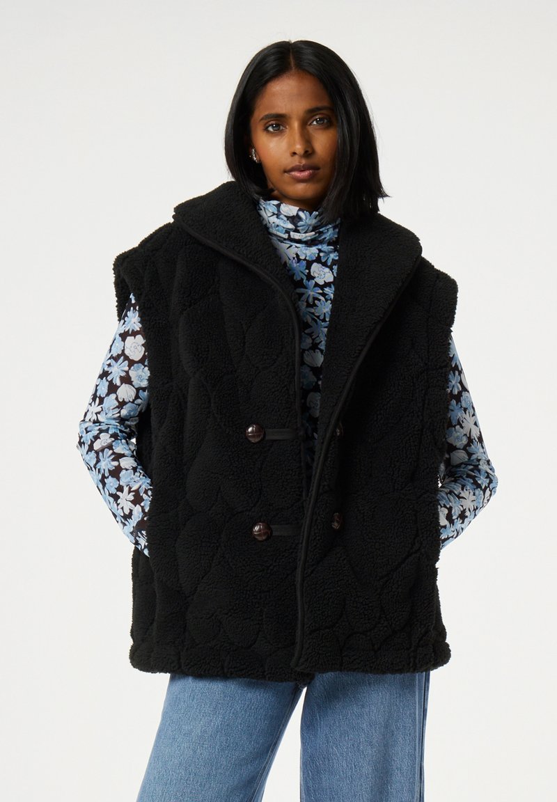Zwarte fleecvest met een hoge kraag, met een gestructureerd ontwerp, knoopsluitingen en voorzakken, gedragen over een bloemige blouse met lange mouwen.