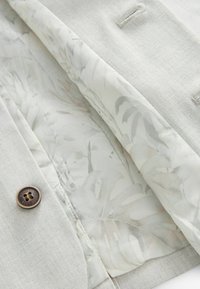 Gros plan d'une veste en tissu beige clair montrant une doublure intérieure à motif floral et un bouton marron à quatre trous avec un texte autour.