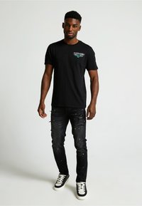 Camiseta de algodón negra con logo en el frente, combinada con jeans de mezclilla negros desgastados que presentan salpicaduras de pintura, y zapatillas negras con acentos blancos.