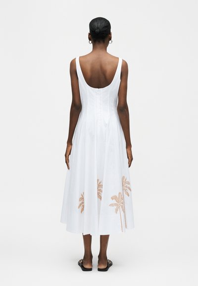 Γυναίκα με σκούρο δέρμα και μαλλιά σε κότσο, ντυμένη με λευκό αμάνικο μεσαίο φόρεμα με καφέ σχέδια φοίνικα στο κάτω μέρος της φούστας, απεικονιζόμενη από πίσω.