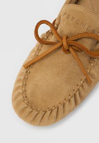 Mocassin en daim beige avec un bout arrondi, présentant un bord cousu et un lacet en cuir noué sur le devant. Marquage discret embossé présent.