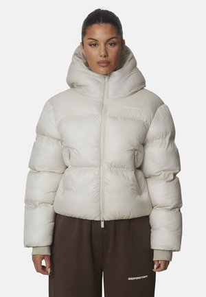 Mujer con chaqueta acolchada color beige claro con cuello alto y pantalones marrón oscuro, de pie sobre un fondo liso.
