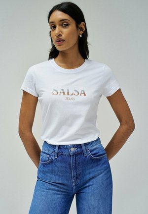 Mujer con camiseta blanca "SALSA JEANS" metida dentro de jeans de mezclilla azul de cintura alta, de pie con las manos detrás de la espalda contra un fondo liso.