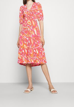 Femme portant une robe courte aux genoux à manches courtes avec un motif abstrait orange, rose et blanc et des sandales blanches à talon bas.