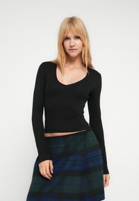 Μαύρο μακρυμάνικο τοπ με V-λαιμόκοψη, κατασκευασμένο από απαλό ύφασμα, εφαρμοστή σιλουέτα, συνδυασμένο με καρό φούστα σε πράσινες και μπλε αποχρώσεις.