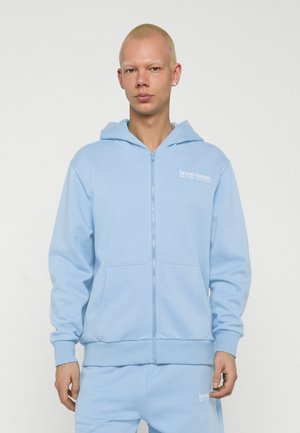 Junge Person mit kurzen blonden Haaren, die einen hellblauen Kapuzenpullover mit Reißverschluss und passende Hosen mit weißen Textlogos trägt.
