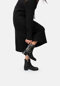 Botas negras hasta el tobillo con un acabado texturizado, featuring dos hebillas de plata, una puntera redonda lisa y un tacón ancho. Cierre de cremallera en la parte posterior.