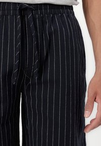 Pantalons rayés bleu marine en tissu léger avec une taille élastique et un cordon de serrage. Présente des rayures blanches verticales pour le détail.