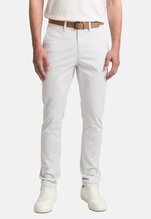 Hellgraue Chinos aus Baumwolle mit schmaler Passform, ausgestattet mit einem vorderen Knopfverschluss, Gürtelschlaufen und Seitentaschen. Kombiniert mit einem weißen Hemd und Turnschuhen.