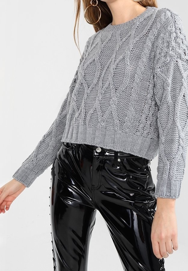 Femme portant un pull court gris en maille torsadée et un pantalon en vinyle noir brillant taille haute avec des détails de laçage sur les côtés.