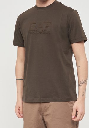 Man draagt een donker olijfgroen EA7 Emporio Armani t-shirt met korte mouwen en beige broek, toont tatoeages op beide onderarmen.