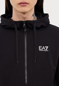 Svart zip-up hoodie med texturerad yta, med huva, dragsko och en EA7 Emporio Armani-logotyp på vänster bröst.