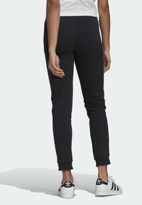 Person som bär svarta slim-fit joggers och vita Adidas-sneakers med svarta ränder, stående på ett ljust grått golv.