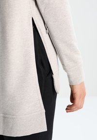 Haut à manches longues gris clair avec une fente latérale, texture douce et poignets côtelés, associé à un pantalon noir avec poches.