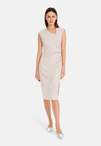 Knee-lange, lichtbeige jurk met kapmouwen, zijsnufjes en een subtiele V-hals. Gecombineerd met glanzende zilveren puntige schoenen.