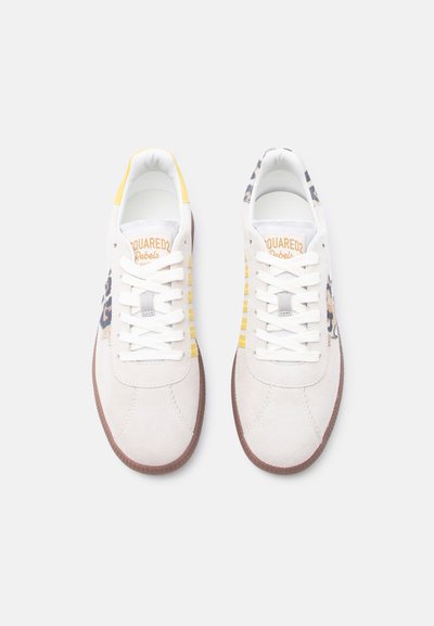 Paire de baskets blanches avec des semelles marron, des bandes jaunes sur les côtés, des accents à imprimé léopard et des lacets blancs sur fond blanc.