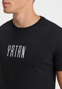 Mladý muž v černém tričku s bílými vertikálními písmeny "YRTRN" vytištěnými na hrudi, sestřiženým v oblasti krku a ramen.