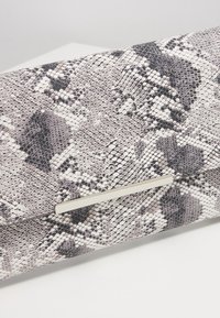 Pochette avec motif peau de serpent texturé gris et noir et une fine fermeture métallique horizontale sur une surface blanche.