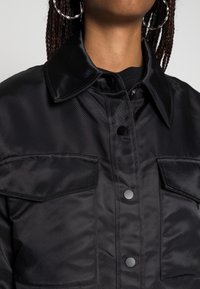 Black satin jacket with a collar, featuring two chest pockets, press-stud fastenings, and a smooth texture.