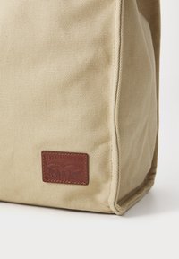 Beige vászontáska, amely texturált felülettel, téglalap alakú dizájnnal és egy barna bőrpárnával rendelkezik, amelyen domborított márkalogó található.