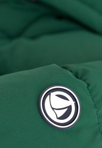 Grønn polstret jakke med en glatt tekstur, med et rundt logo-merke i svart og hvitt på venstre side.