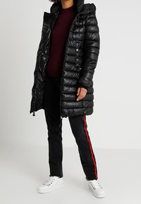 Doudoune noire avec capuche et finition brillante, arborant un design matelassé et des boutons pression. Portée par-dessus un pull bordeaux et un pantalon noir.
