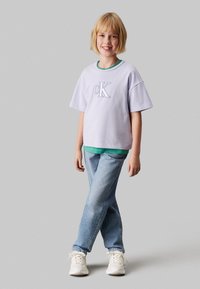 Camiseta de color púrpura claro con logo bordado "CK", superpuesta sobre una camiseta verde; combinada con jeans azules holgados y zapatillas blancas.