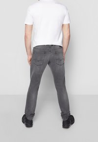 Homme debout de dos, portant une chemise blanche à manches courtes, un jean gris en denim et des chaussures noires sur un fond uni.