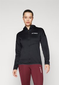 Čierna mikina na zips s kapucňou, na prednej strane s malým bielym logom TERREX, vyrobená z hladkej, elastickej textílie.