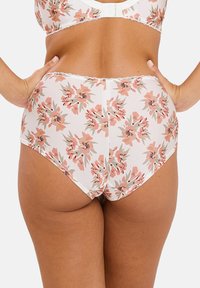 Femme portant des sous-vêtements taille haute à motifs floraux blancs et un soutien-gorge assorti, se tenant avec les mains sur les hanches, vue de dos sur fond uni.
