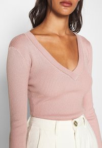 Geribbeld, lichtroze top met lange mouwen en een diepe V-hals, gemaakt van textiel met structuur, gecombineerd met hooggeritste, lichtgekleurde broek met een knoop.