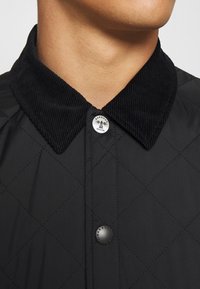 Veste noire matelassée avec un col en velours côtelé texturé, dotée d'une fermeture à boutons-pression et d'un détail métallique de marque.