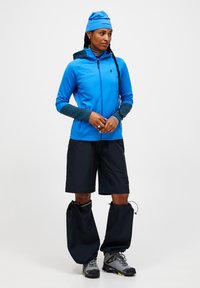 Blauwe zip-up jas met grijze accenten, gecombineerd met zwarte shorts. Bevat grote gaiters en grijze wandelschoenen. Stof lijkt lichtgewicht en ademend.