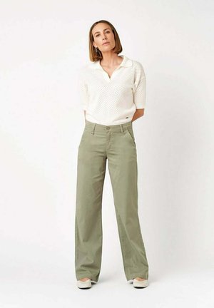 Donna con capelli lisci fino alle spalle indossa un top bianco a maglia a maniche corte e pantaloni verde oliva a vita alta e gamba larga, in piedi davanti a uno sfondo bianco.