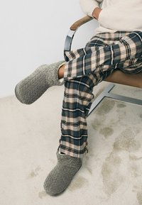 Grå teksturerede slippers med et mønstret overflade, parret med ternede pyjamasbukser i marineblå, beige og hvid. Sidder på en stol.