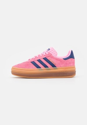 Roze Adidas Gazelle sneaker met marineblauwe strepen en dikke, gelaagde gumzool op een witte achtergrond, zijaanzicht.