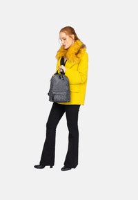 Cappotto giallo con colletto in pelliccia, top nero, pantaloni neri a campana e stivali neri; tiene uno zainetto grigio piccolo con motivo a logo.