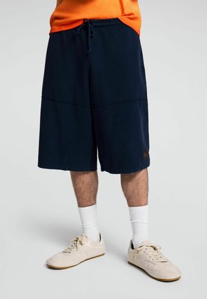 Persona che indossa pantaloncini blu navy testurizzati fino al ginocchio, calzini bianchi, scarpe da ginnastica beige e una maglietta arancione corta in vita.
