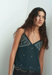 Top in teal scuro con dettagli ricamati in un motivo geometrico, spalline delicate e un fit rilassato con una texture leggermente trasparente.