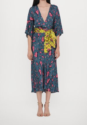 Femme portant une robe portefeuille bleue à fleurs roses avec une large ceinture fleurie jaune nouée en nœud, associée à des talons beiges à lanières.