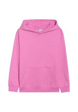 Młoda osoba z długimi brązowymi włosami w różowej bluzie z kapturem, beżowych spodniach i szarych sneakersach, stoi z rękami w kieszeniach bluzy.