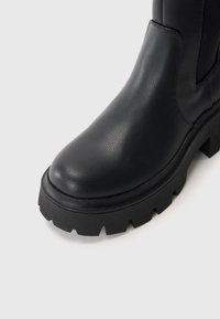 Crne kožne čizme do gležnja s zaobljenim vrhom, debelim potplatom s režnjima i glatkom teksturom. Ima minimalno šivanje i elegantan dizajn.