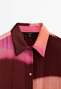 Chemise à col boutonné avec un motif dégradé dans des nuances de bordeaux, rose et pêche, montrant les boutons du haut et une partie du col.