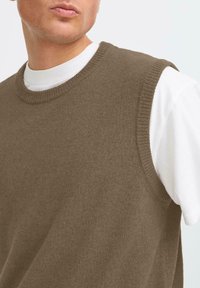 Suéter sin mangas de punto marrón con cuello acanalado, llevado sobre una camiseta blanca de cuello redondo. La textura parece suave y fina.